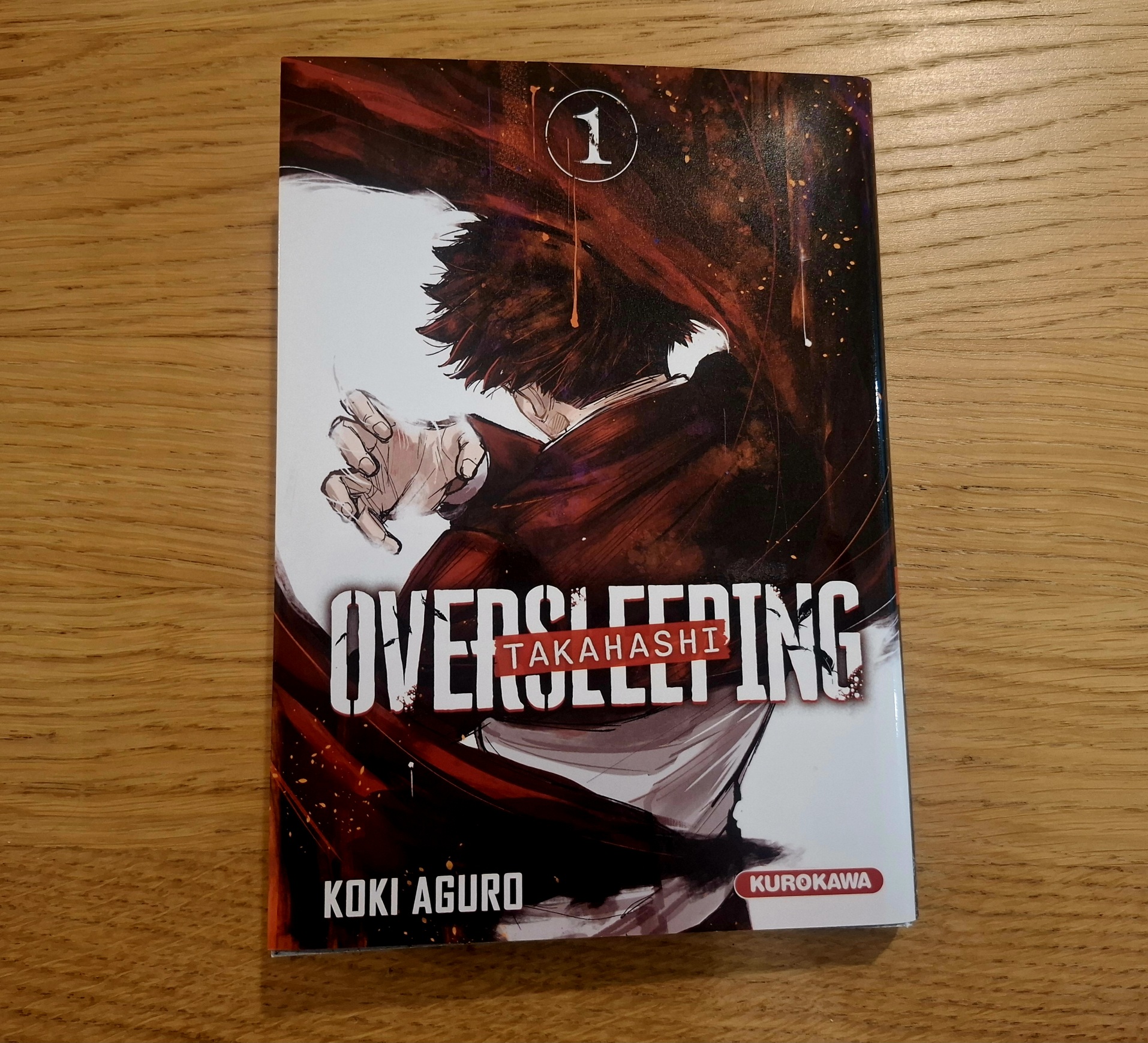 Le tome 1 du manga Oversleeping Takahashi posé sur une table, couleurs dark à dominance de noir, rouge et blanc